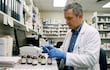 Hombre en bata blanca y guantes azules manipula medicamentos en una farmacia, rodeado de frascos y documentos en la mesa.