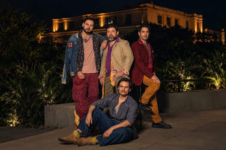 Cuatro hombres posan en un evento al aire libre, vestidos con chaquetas y pantalones coloridos, en un ambiente iluminado y relajado.