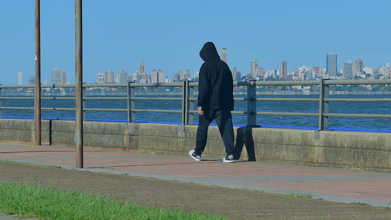 Persona caminando sola por la costanera, vestida con sudadera negra y pantalones oscuros, ciudad moderna al fondo.