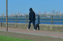 Persona caminando sola por la costanera, vestida con sudadera negra y pantalones oscuros, ciudad moderna al fondo.