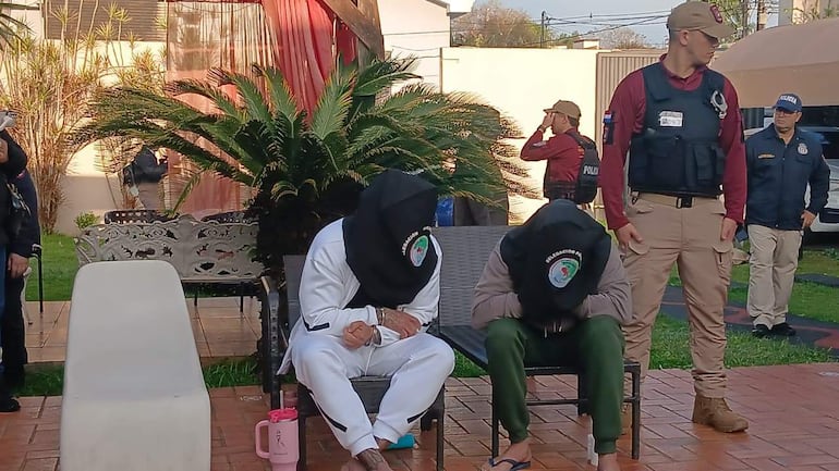 Dos personas con capuchas negras, una en conjunto deportivo blanco y otra en ropa gris, sentadas mientras policías las observan.