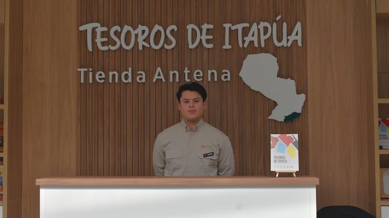 Lucas, joven con camisa clara, atiende en mostrador de Tienda Antena 'Tesoros de Itapúa', rodeado de decoración de madera.