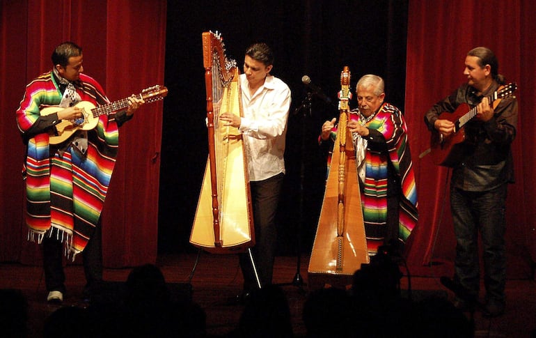 Cuatro hombres en ponchos multicolores tocan arpa y guitarras en un teatro con telón rojo de fondo.