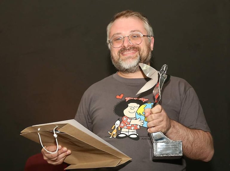Hombre con camiseta de dibujos animados sonríe al recibir un trofeo y una bolsa de papel en un fondo oscuro.