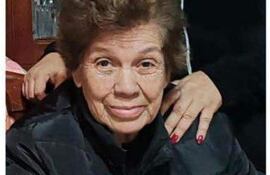 Mujer de 84 años con cabello rizado y chaqueta negra, sentada en un ambiente acogedor, con mano de otra persona sobre su hombro.