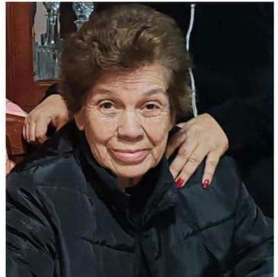 Mujer de 84 años con cabello rizado y chaqueta negra, sentada en un ambiente acogedor, con mano de otra persona sobre su hombro.