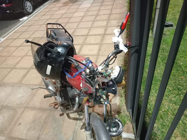 Motocicleta roja parcialmente desmantelada en una calle, con un casco negro sobre el manillar, rodeada de césped y pavimento.