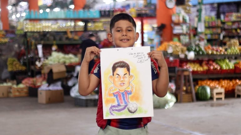Niño sonriente en camiseta de franjas rojas y azules sostiene un dibujo de un futbolista en un mercado lleno de color.