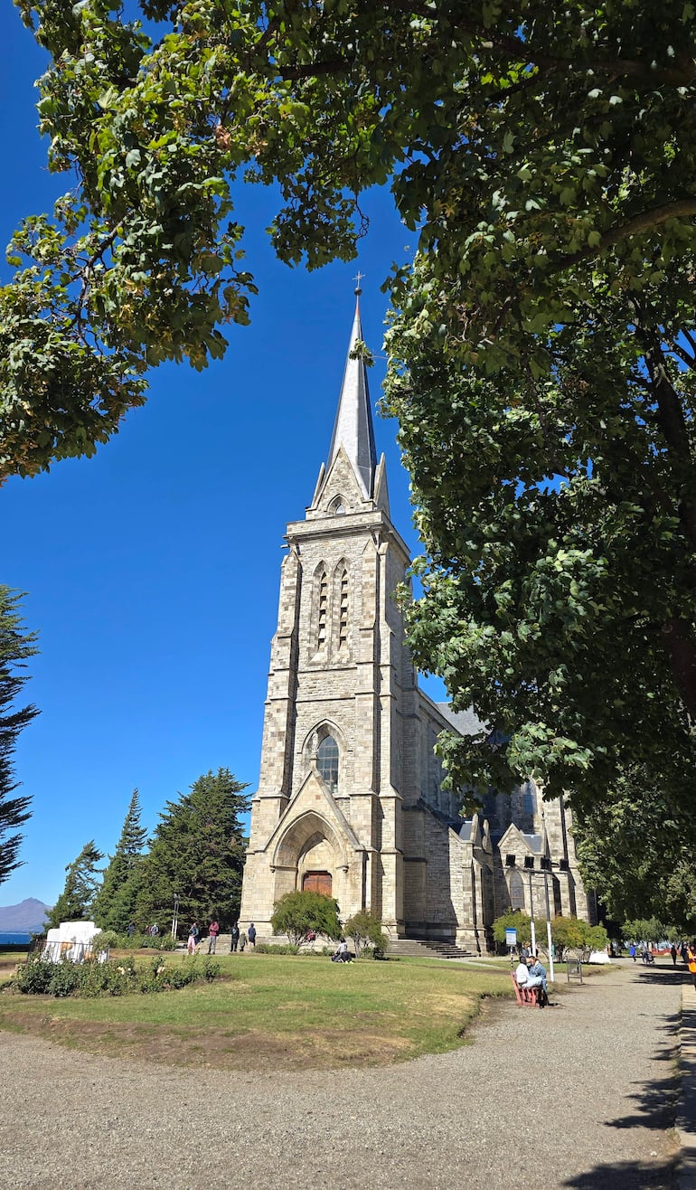 Iglesia gótica con campanario rodeada de árboles, personas caminan y se sientan en bancos en un entorno soleado.