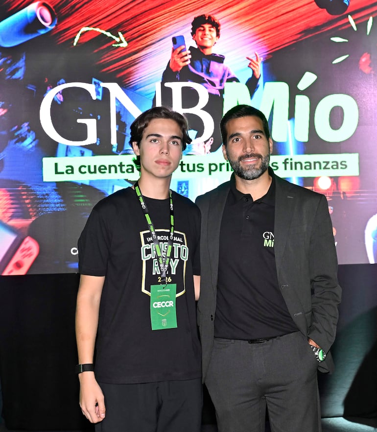 Hombre en traje negro presenta mientras joven con camiseta negra 'Cristo Rey' escucha atentamente, fondo con logo GNB Mío.