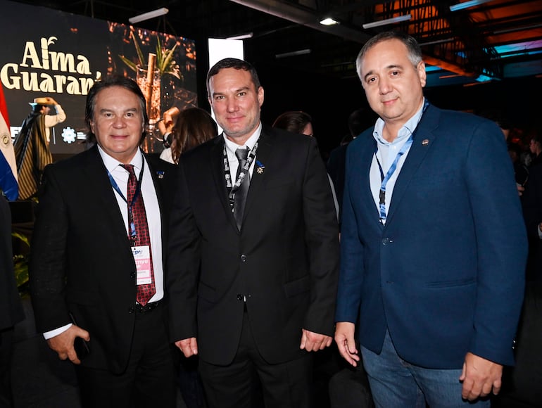 Tres hombres de pie en evento: uno con camisa blanca y corbata roja, otro en traje negro, el tercero con abrigo azul, fondo oscuro con pantalla.