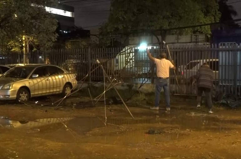 Dos hombres, uno de pie y otro arrodillado, trabajando en estructuras en un ambiente improvisado, con un automóvil plateado al fondo.