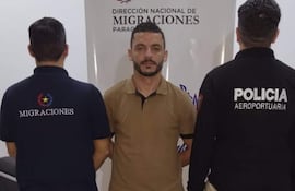 Hombre de camiseta beige al frente, dos agentes de espaldas con camisetas de 'MIGRACIONES' y 'POLICÍA AEROPORTUARIA' en oficina.