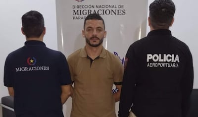 Hombre de camiseta beige al frente, dos agentes de espaldas con camisetas de 'MIGRACIONES' y 'POLICÍA AEROPORTUARIA' en oficina.