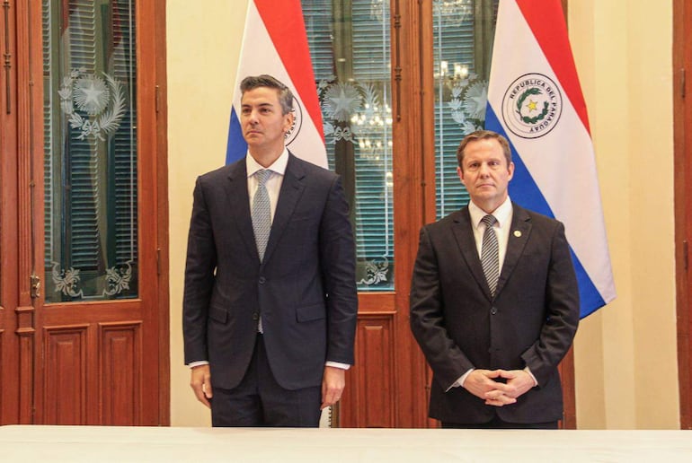 Alberto Joaquín Martínez Simón y Alberto Jiménez Rolón en trajes formales, serios, en recinto oficial con banderas paraguayas al fondo.