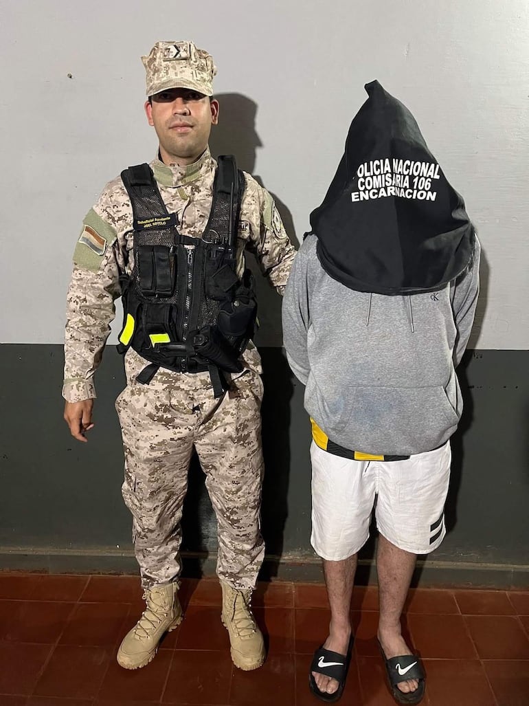 Oficial de Policía Nacional en uniforme camuflado, custodia a joven con capucha negra y sudadera gris en comisaría.