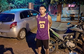 Joven de 18 años con camiseta morada de los Lakers junto a motocicleta robada y automóvil plateado en un entorno urbano.