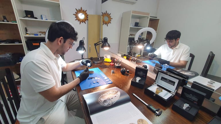 Hombre con guantes negros y camiseta blanca, concentrado en mesa llena de relojes y herramientas, en ambiente de trabajo.