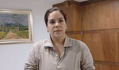 Johanna Ortega en camisa clara, reflexionando en oficina con pared de madera y pintura de paisaje al fondo.