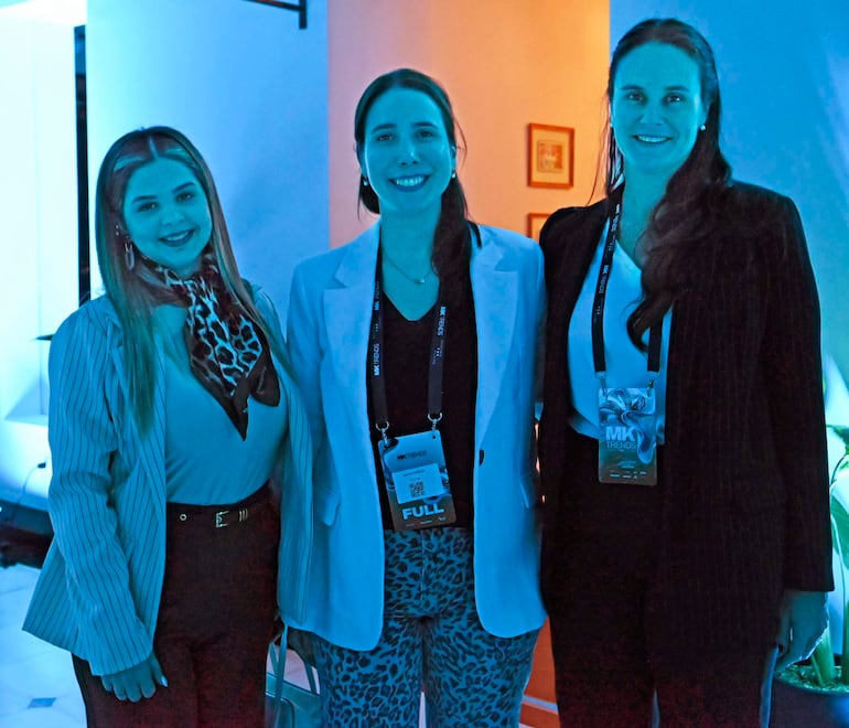 Tres mujeres sonrientes, con diferentes chaquetas, posan en un entorno iluminado con luces azules.