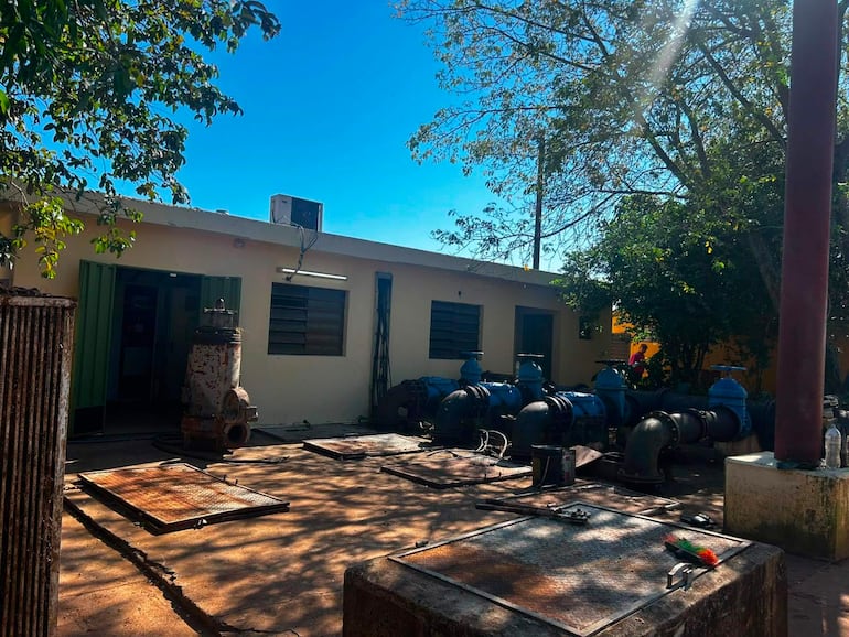 Edificio de una planta con puerta verde y tuberías azules, rodeado de árboles y suelo de cemento, sin personas presentes.