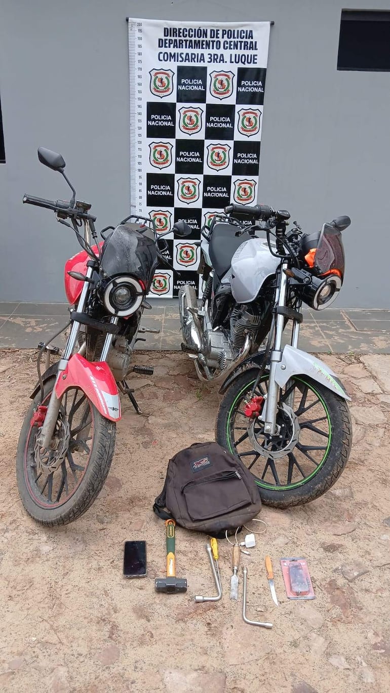 Dos motocicletas, una roja y una blanca, expuestas en un entorno policial, rodeadas de herramientas y objetos personales en un espacio al aire libre.