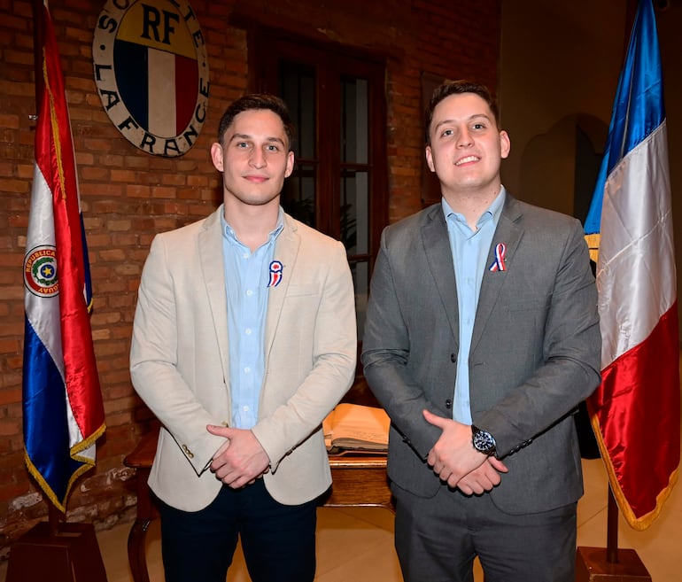 Dos hombres en trajes formales, uno con saco claro y el otro con traje gris, posan ante banderas de Paraguay y Francia.