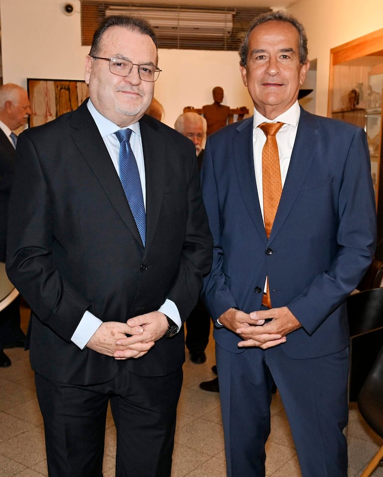 Dos hombres en trajes oscuros con corbatas diferentes, en un salón de eventos, interactuando con posturas serias.
