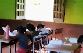 Cuatro niños, dos con cabello recogido y camisas de colores, estudian en escritorios de metal en un aula con paredes verdes y luz natural.