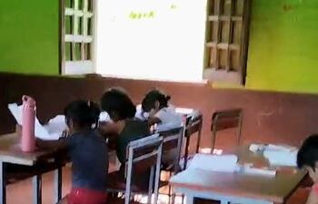 Cuatro niños, dos con cabello recogido y camisas de colores, estudian en escritorios de metal en un aula con paredes verdes y luz natural.