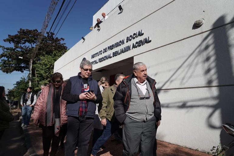 Isaías Fretes con teléfono en mano, rodeado de personas con abrigos, frente al IPS Hospital Regional Benjamín Aceval bajo un cielo soleado.