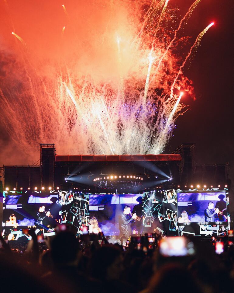 Ricardo Arjona en el escenario con artistas, iluminado por fuegos artificiales rojos y azules, mientras la audiencia graba el momento.