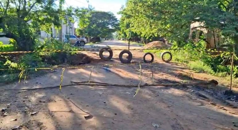 Calle residencial bloqueada con neumáticos y cintas de advertencia amarillas; vegetación a los lados.
