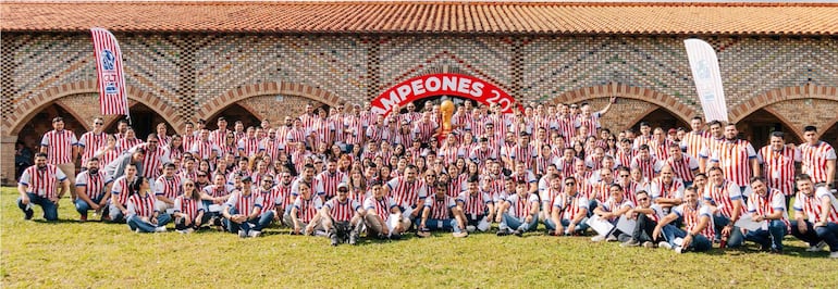 Ciento de personas sonrientes en camisetas a rayas rojas y blancas posan junto a un trofeo, con una banderola que dice 'CAMPEONES 2022' en el fondo.