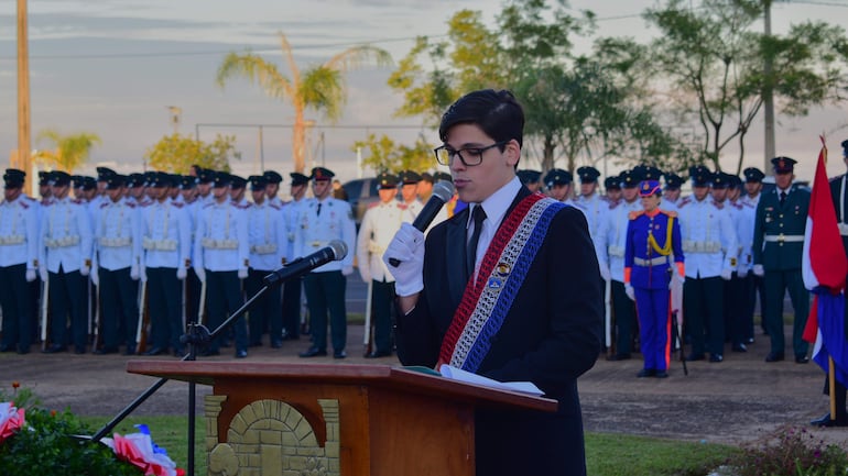 Estudiante del colegio Bautista en traje negro con banda nacional, hablando frente a soldados en uniforme blanco en un acto formal al aire libre.