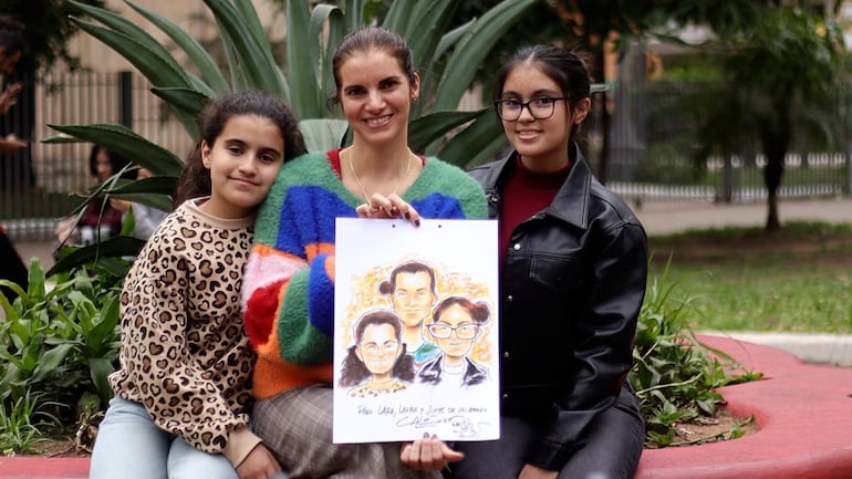 Niña con suéter animal sonriente, mujer con suéter multicolor sosteniendo dibujo, joven con gafas y suéter negro, todo rodeado de vegetación.