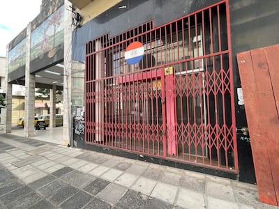 Fachada de local con rejas rojas y bandera paraguaya, sin personas visibles en el entorno urbano.