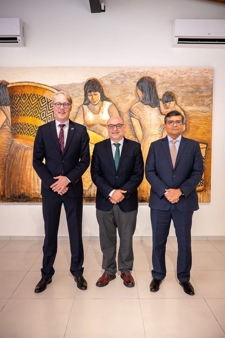 Tres hombres en trajes oscuros frente a una pintura decorativa, con uno usando gafas y otro con corbata verde.