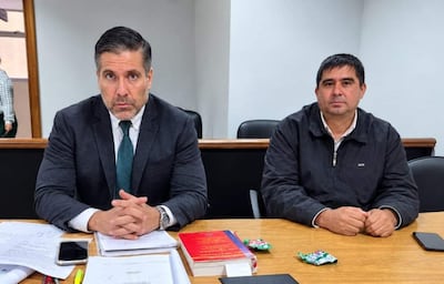 Víctor Garay Olmedo en traje oscuro con corbata verde, discutendo con un colega de chaqueta oscura en una mesa de madera.
