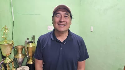Hombre sonriendo con camiseta azul y gorra, rodeado de trofeos dorados y plateados en un ambiente interior verde.