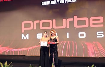 Mujeres felices en el escenario, una con trofeo y otra con documento, fondo decorado con vegetación y logo de prourbe.