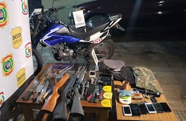 Mesa con armas, teléfonos y evidencia del operativo; motocicleta Kenton azul al fondo, con logos de la Policía Nacional de Paraguay.