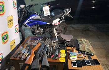 Mesa con armas, teléfonos y evidencia del operativo; motocicleta Kenton azul al fondo, con logos de la Policía Nacional de Paraguay.
