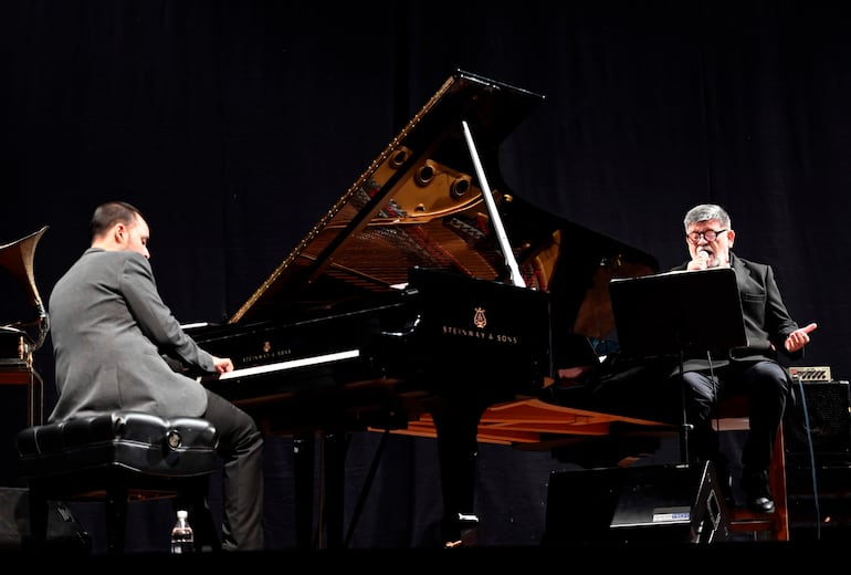 Pianista de traje gris oscuro toca un piano Steinway mientras un hombre con chaqueta negra canta con micrófono en mano.