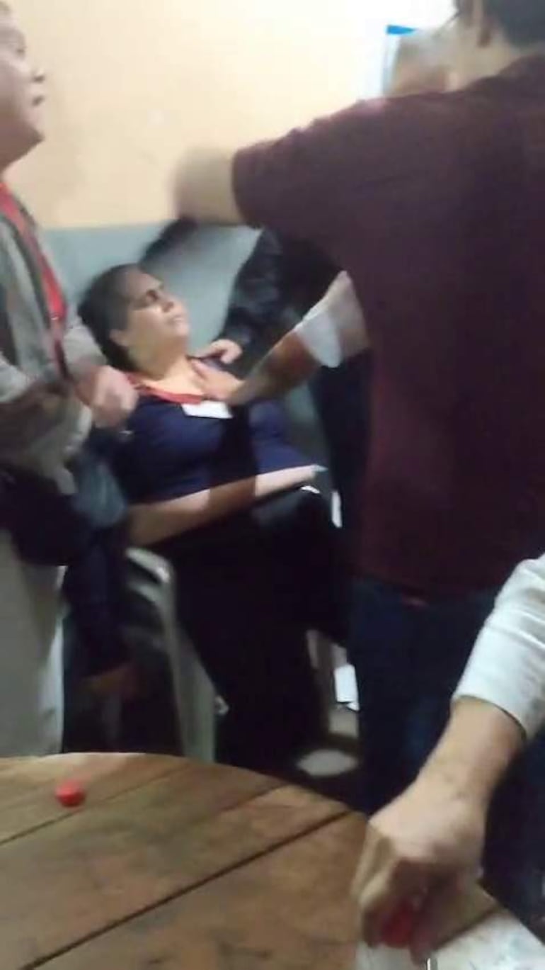 Mujer con cabello largo y oscuro sentada, mostrando malestar, rodeada de tres personas en un ambiente tenso.