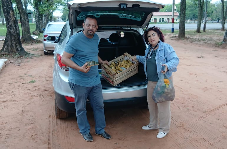 Hombre sostiene torta de banana y mujer con bolsa plástica y caja de frutas en un entorno natural.