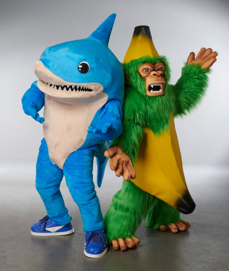 Personajes disfrazados: tiburón azul inclinado hacia adelante y gorila verde con una banana, ambos de pie en un ambiente neutro.