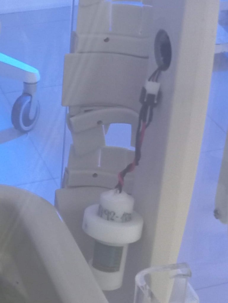 Dispositivo médico con cuerpo blanco y cableado, en ambiente iluminado con tonos azulados, dentro de un hospital.