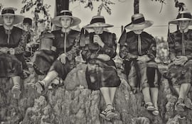 Seis mujeres menonitas sentadas sobre rocas, vestidas de oscuro y con sombreros amplios, en un entorno natural tranquilo.
