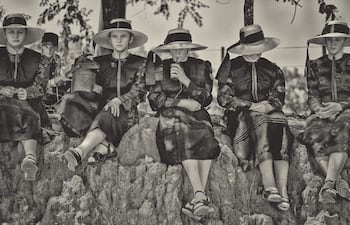 Seis mujeres menonitas sentadas sobre rocas, vestidas de oscuro y con sombreros amplios, en un entorno natural tranquilo.